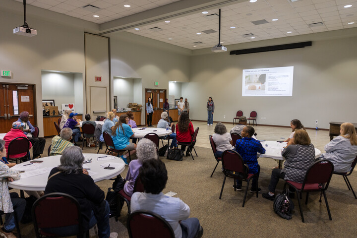 Participants seated around tables listen to a presentation in a community hall.