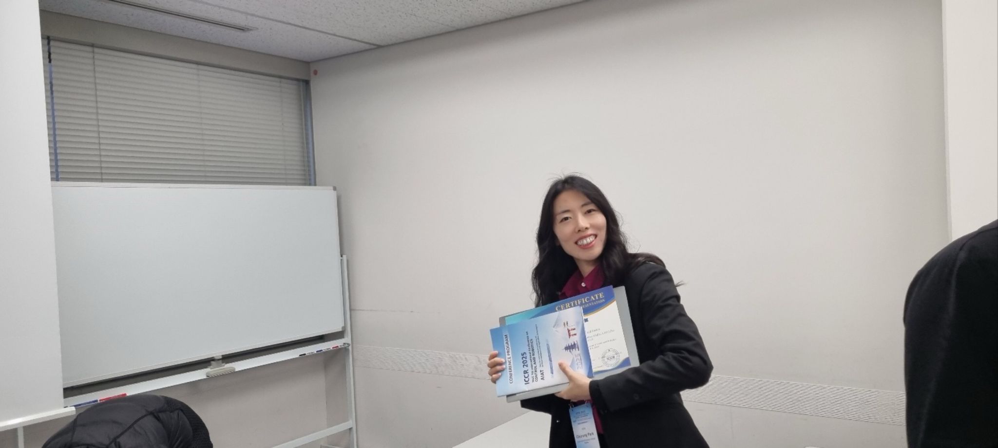 A presenter smiles while holding a certificate in a conference room.
