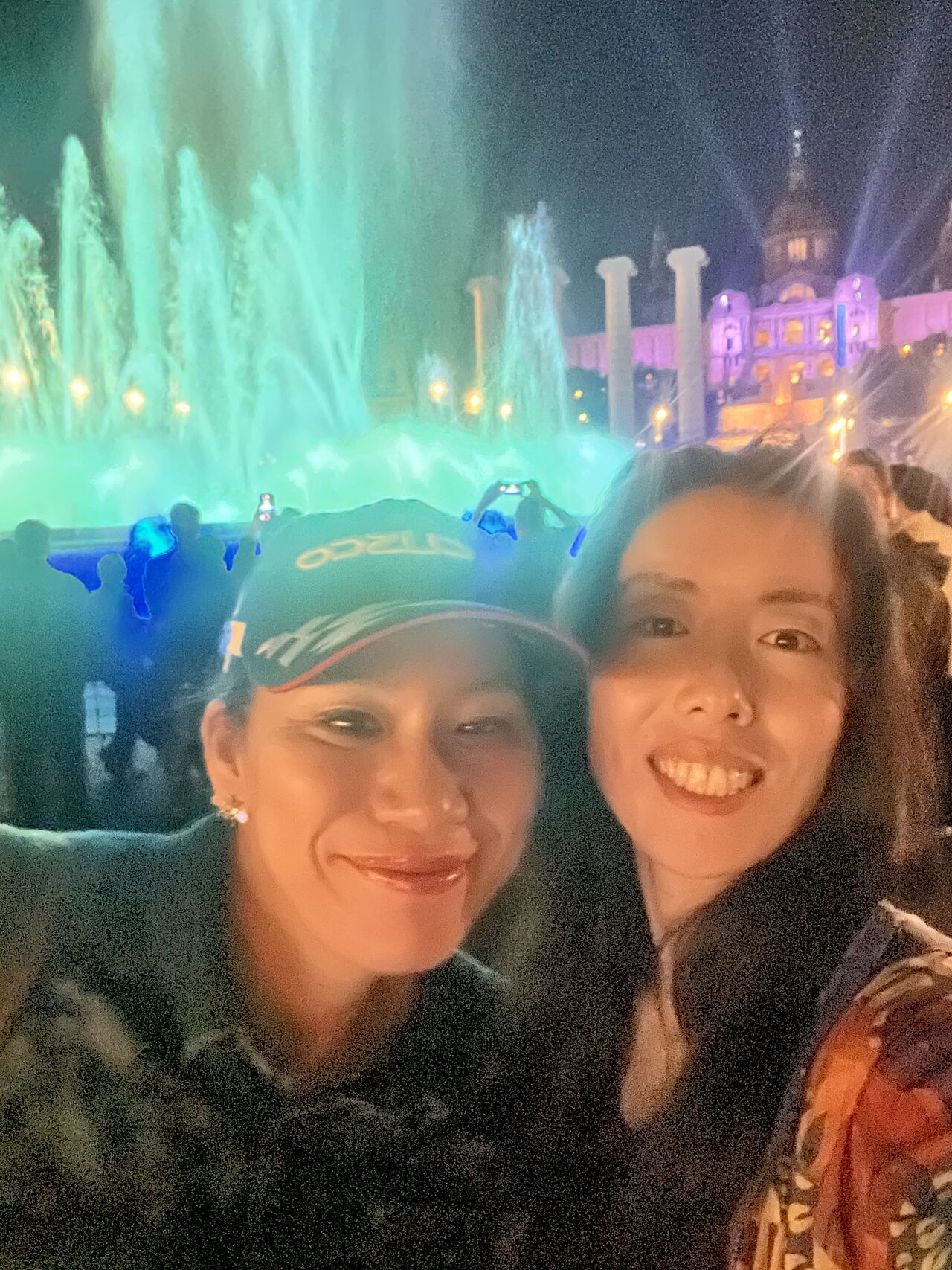 Two attendees take a photograph beside illuminated fountains at night.