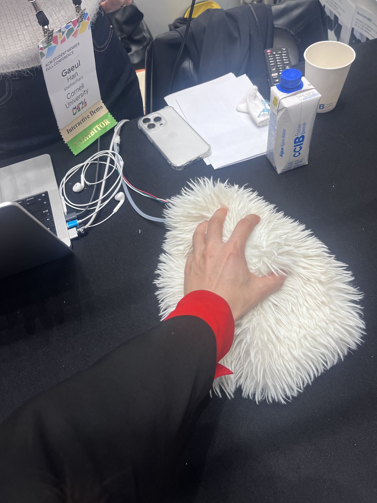 A hand touches a white furry robotic device on a demonstration table.