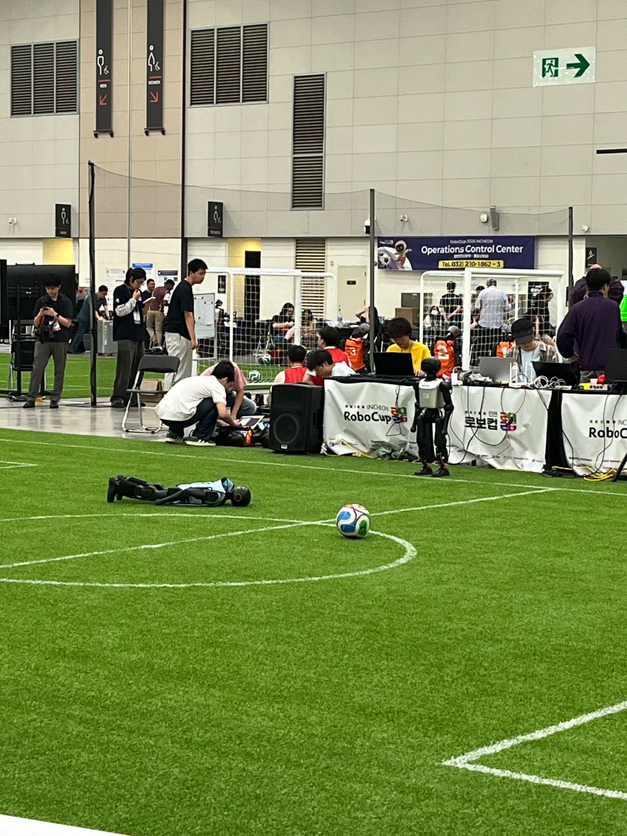 Robots and participants occupy a green indoor soccer field.