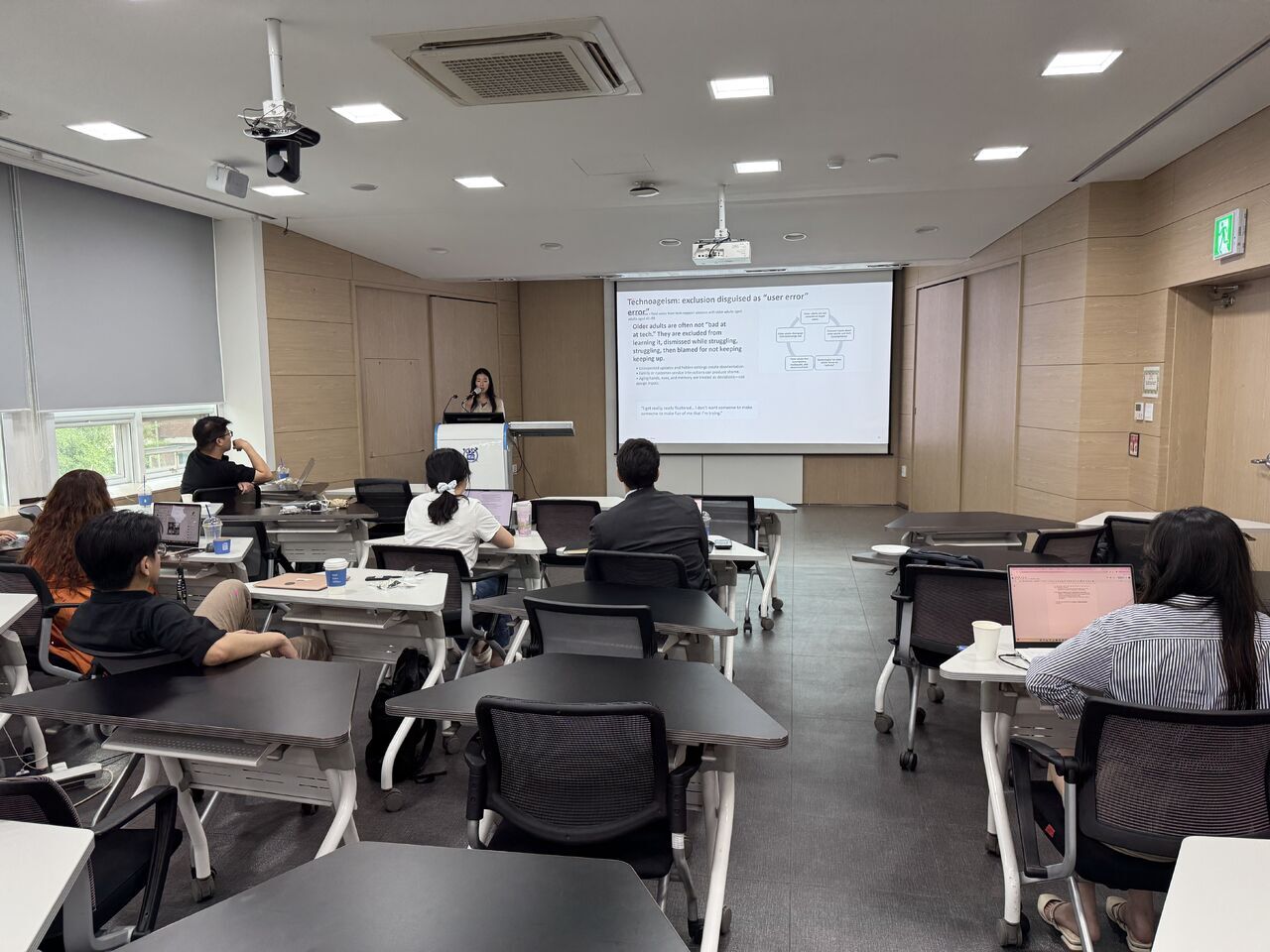 Audience members sit at classroom desks facing a presenter and screen.