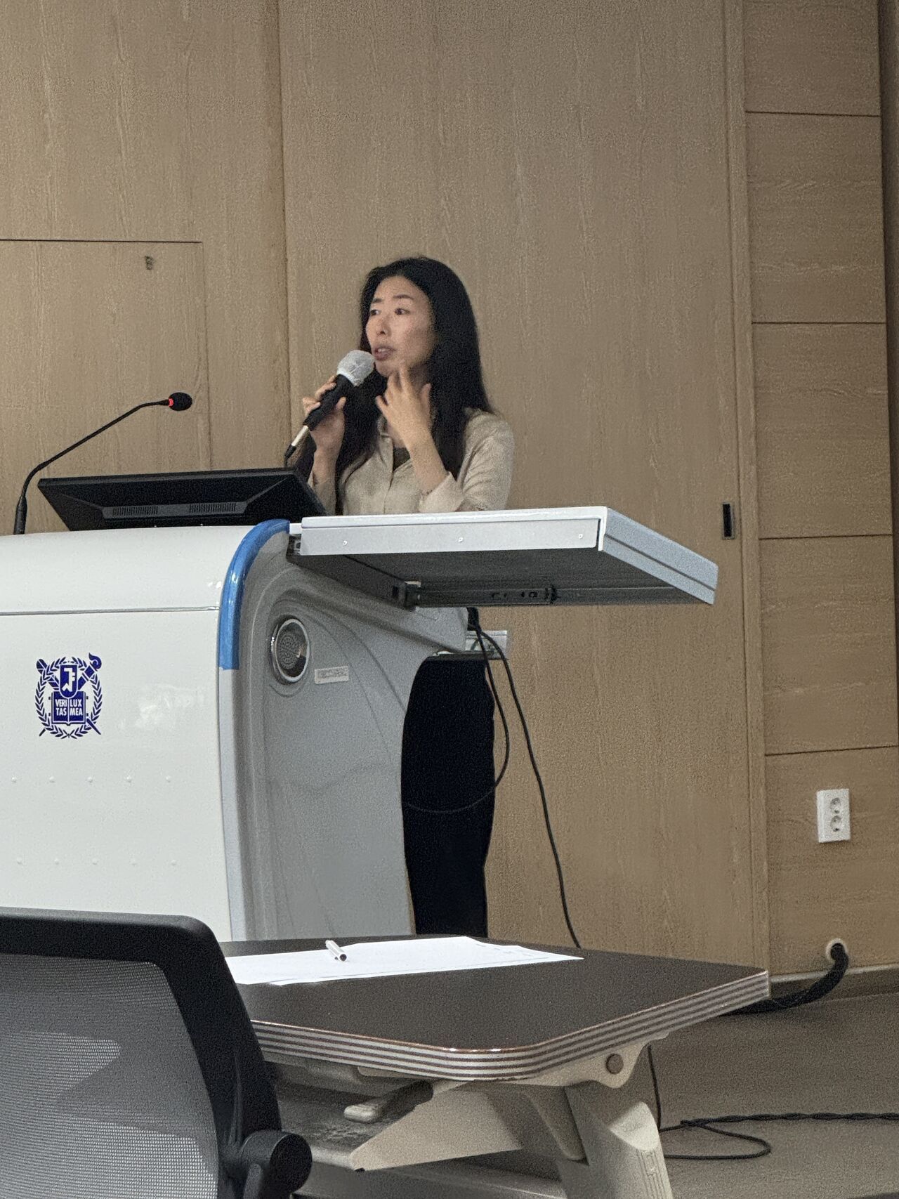 A presenter holds a microphone behind a university lectern.