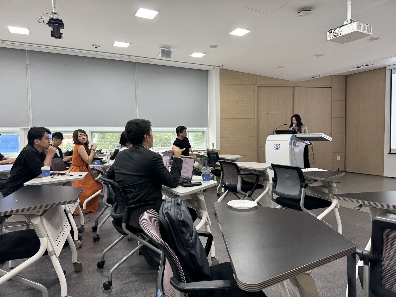 A wide view of classroom participants, desks, and the presenter.