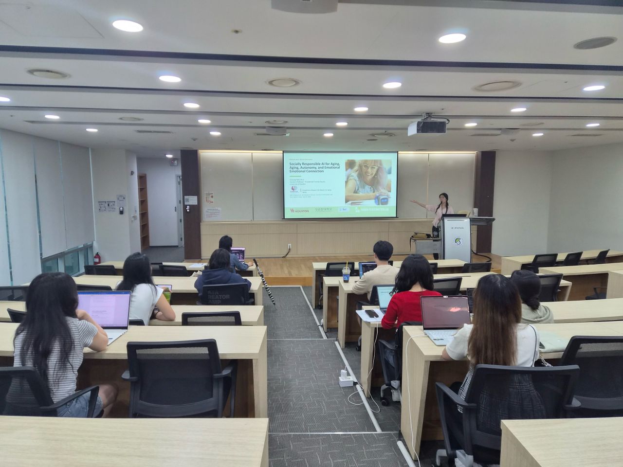 An audience sits in a lecture room facing a presenter and screen.