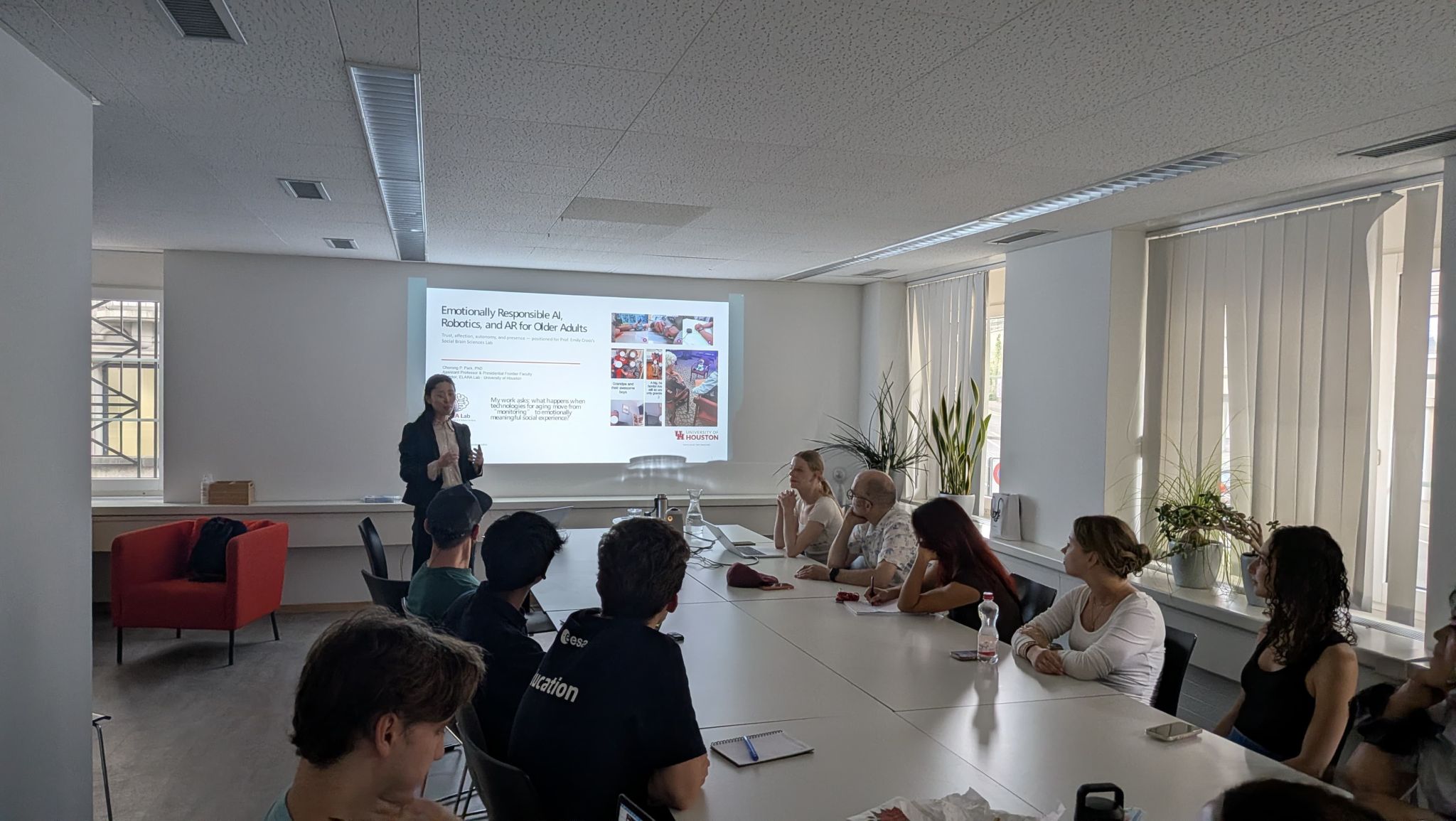 A research group watches a presentation beside tall windows.
