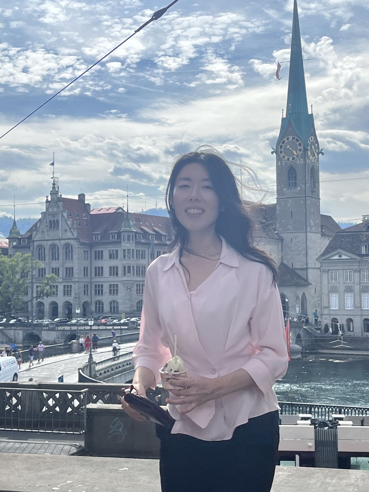 A visitor stands beside the river with Zurich buildings and a church spire behind her.