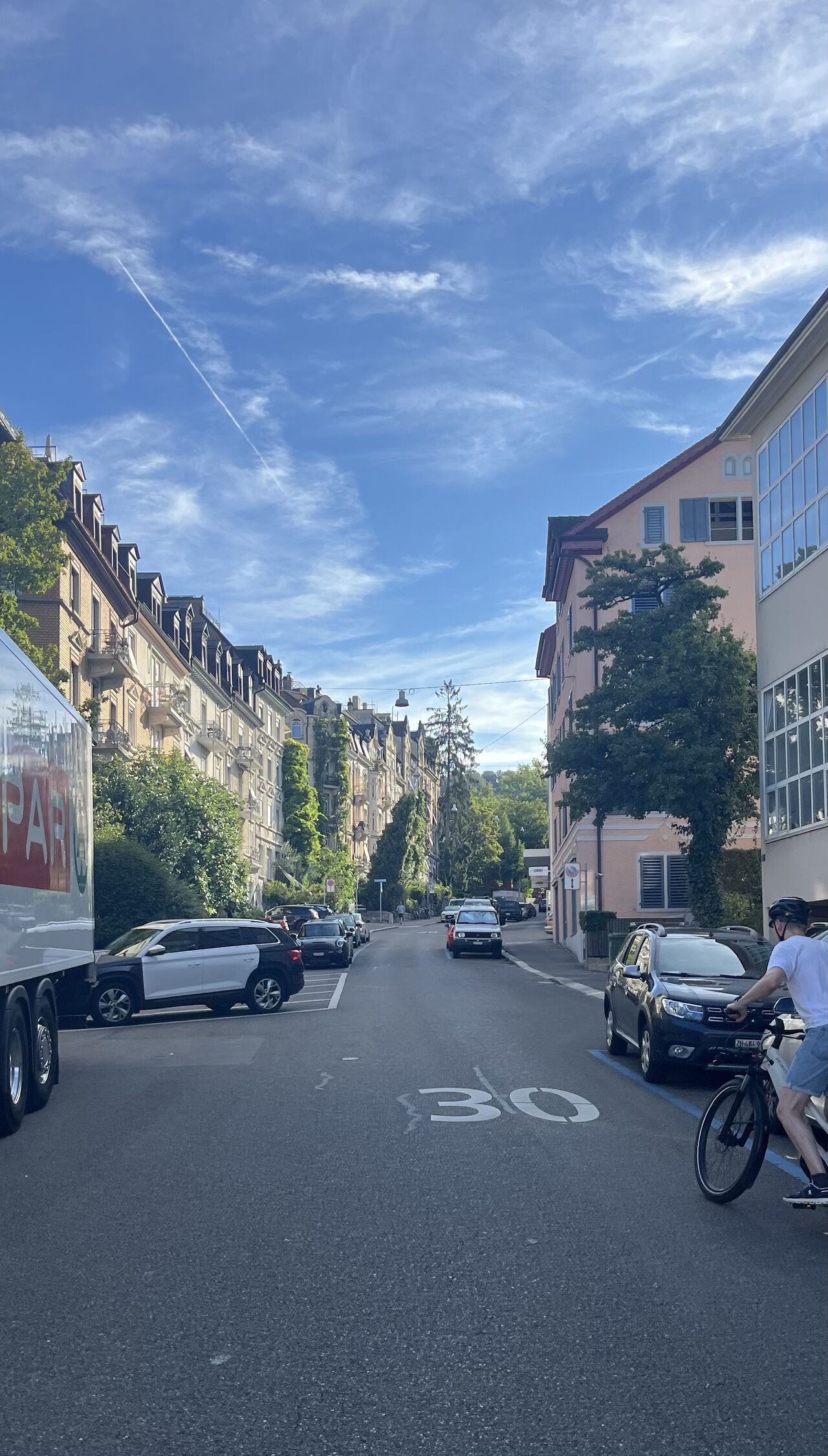 A sunlit urban street lined with buildings and parked vehicles.