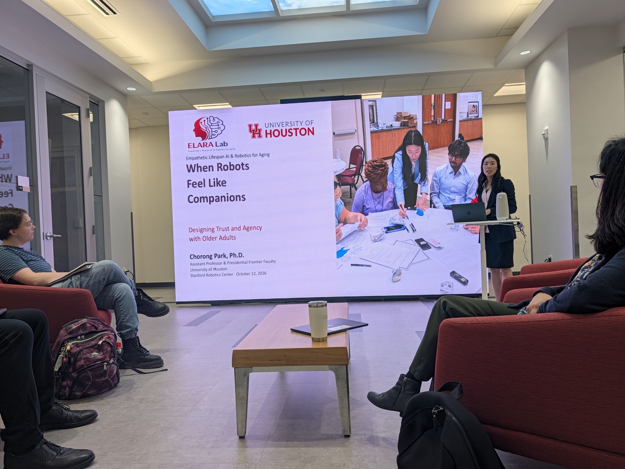 A speaker stands beside a title slide about companion robots, trust, and agency while an audience sits in red chairs.