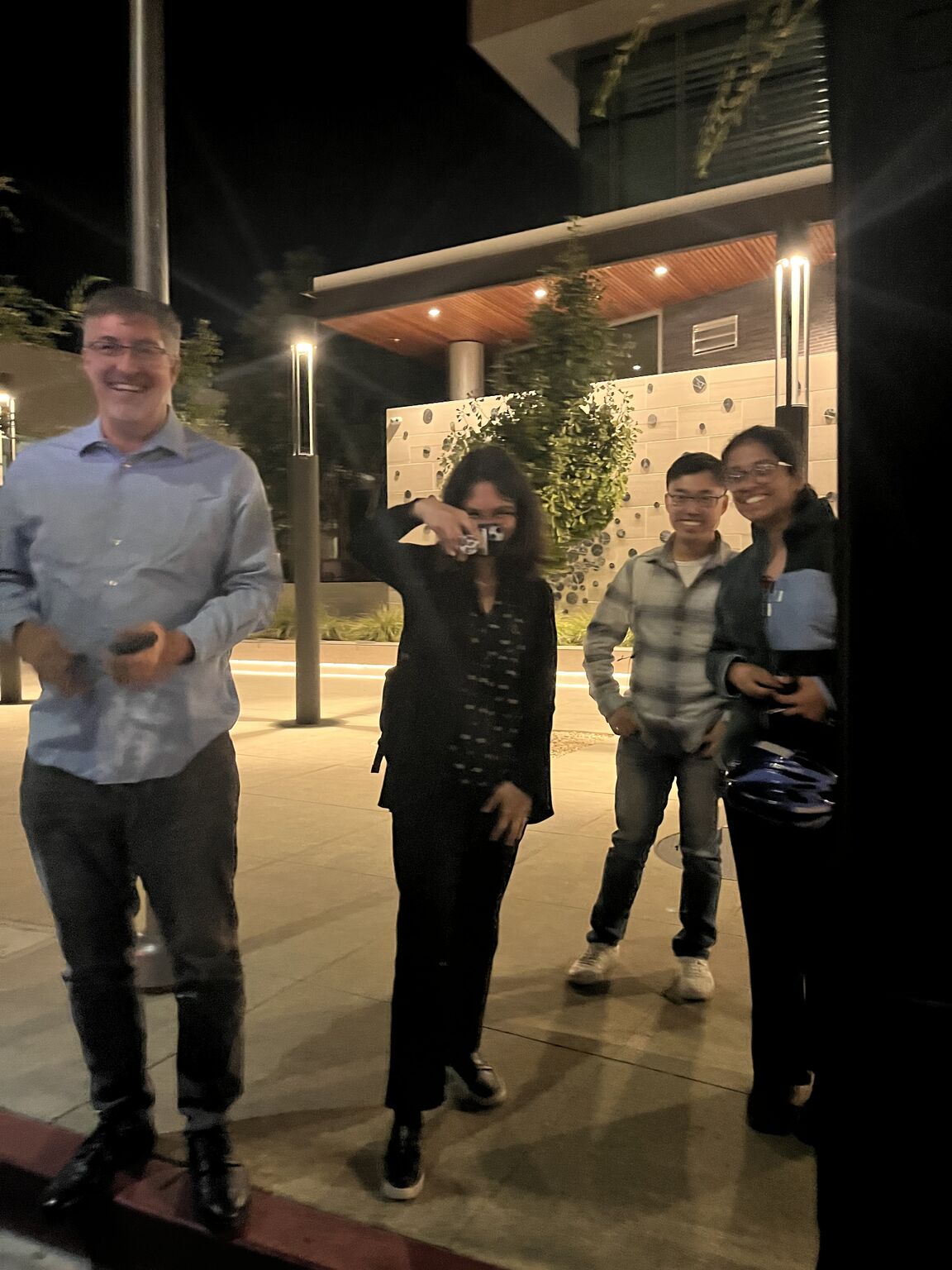 Four people stand outside a building at night, with one holding up a phone.