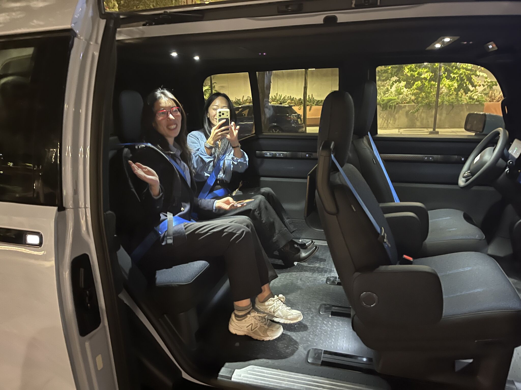 Two people sit inside a vehicle with blue seat belts, one holding up a phone.