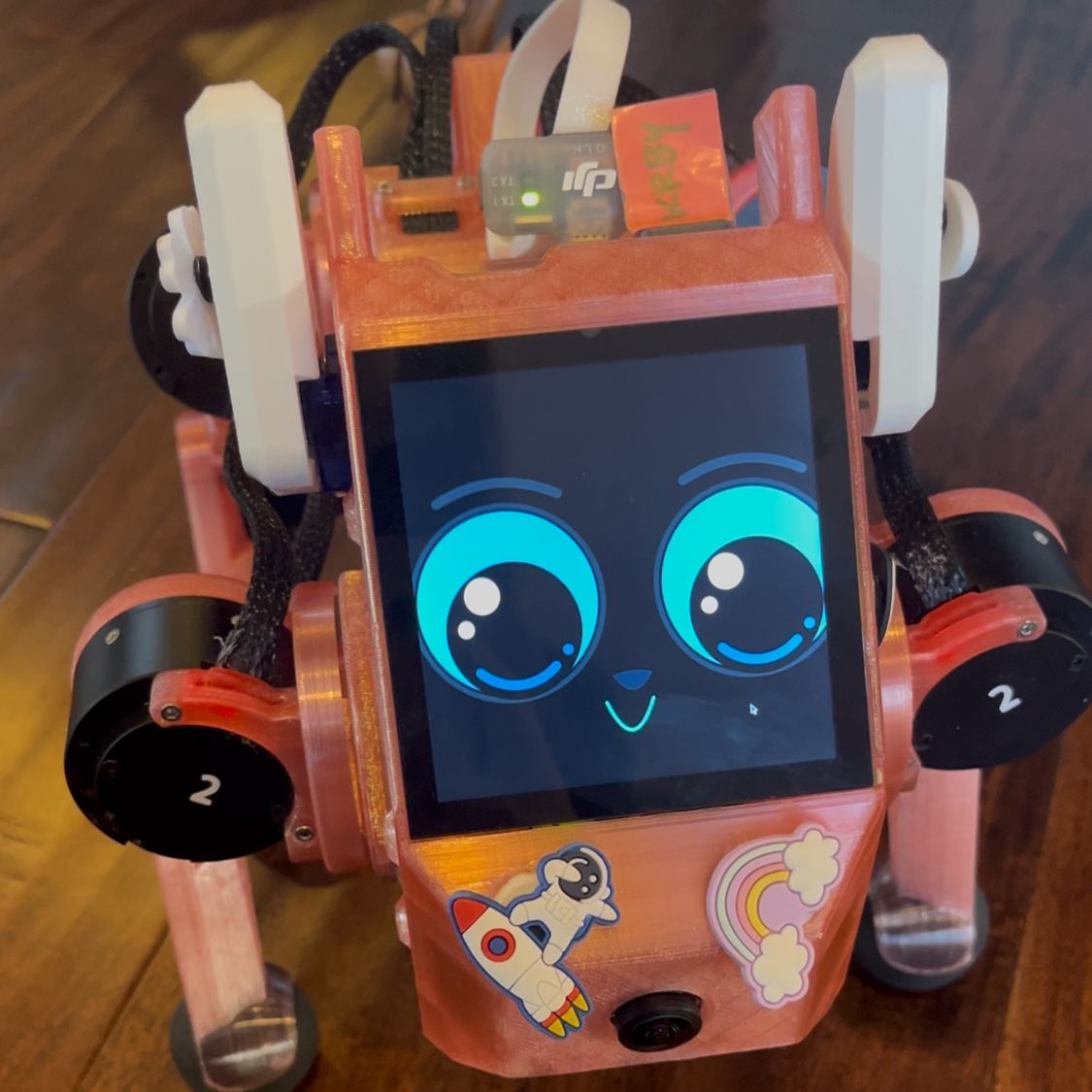 A small pink four-legged robot has a screen face with blue eyes and colorful stickers on its body.