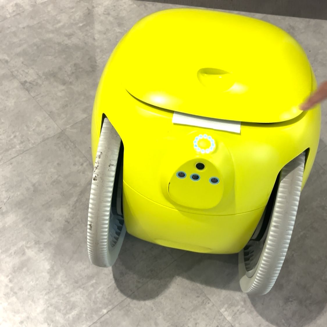 A bright yellow wheeled robot with a top lid stands on a gray floor.