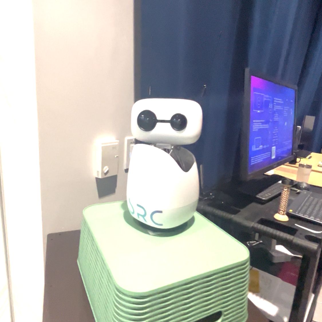 A white robot with two dark circular eyes sits on a green platform beside a computer monitor.