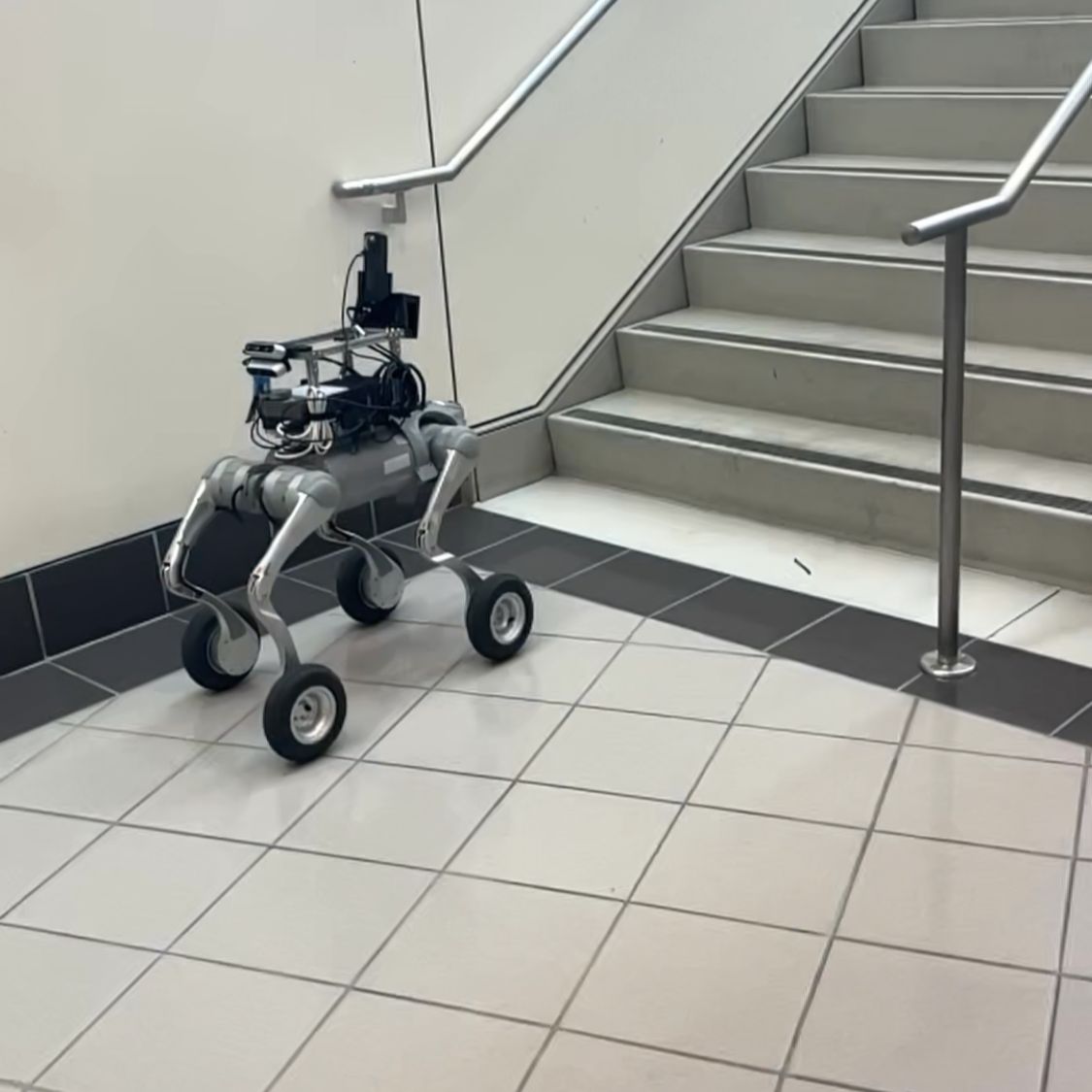 A four-legged robot with wheels and mounted equipment stands at the foot of an indoor staircase.