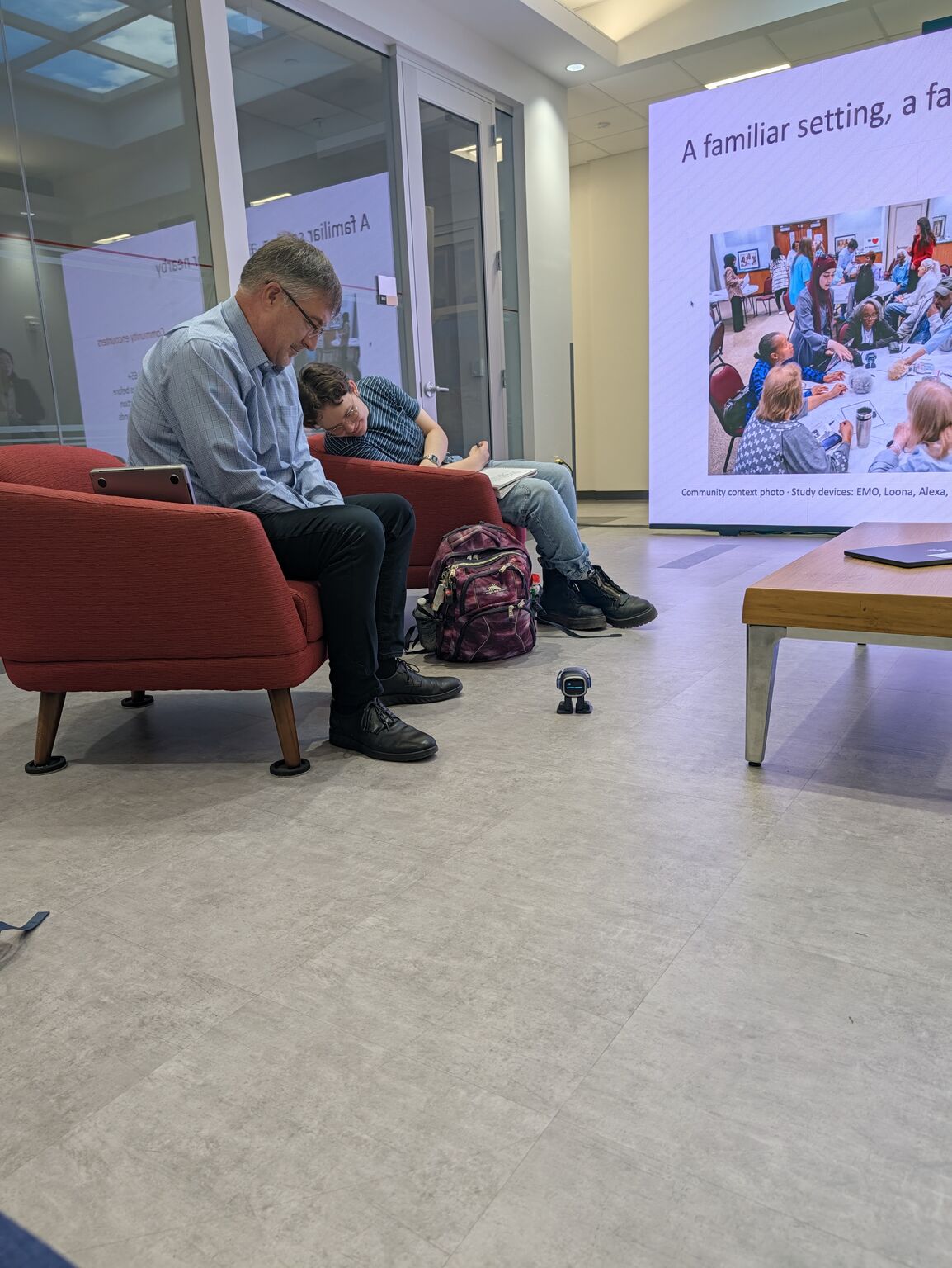 Two seated attendees look down toward a small robot beside a presentation screen.