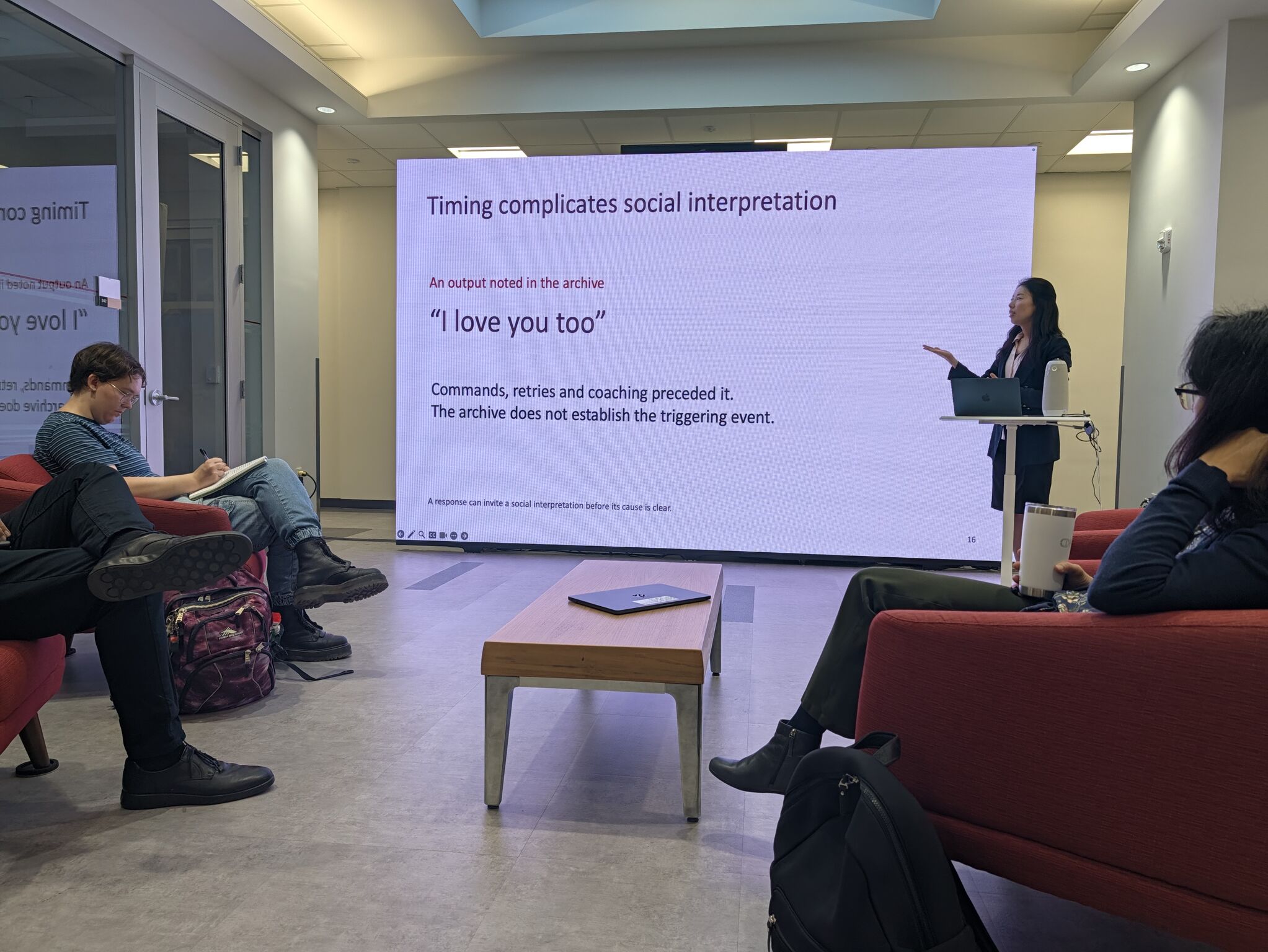 A speaker gestures beside a slide about how timing complicates the social interpretation of robot responses.