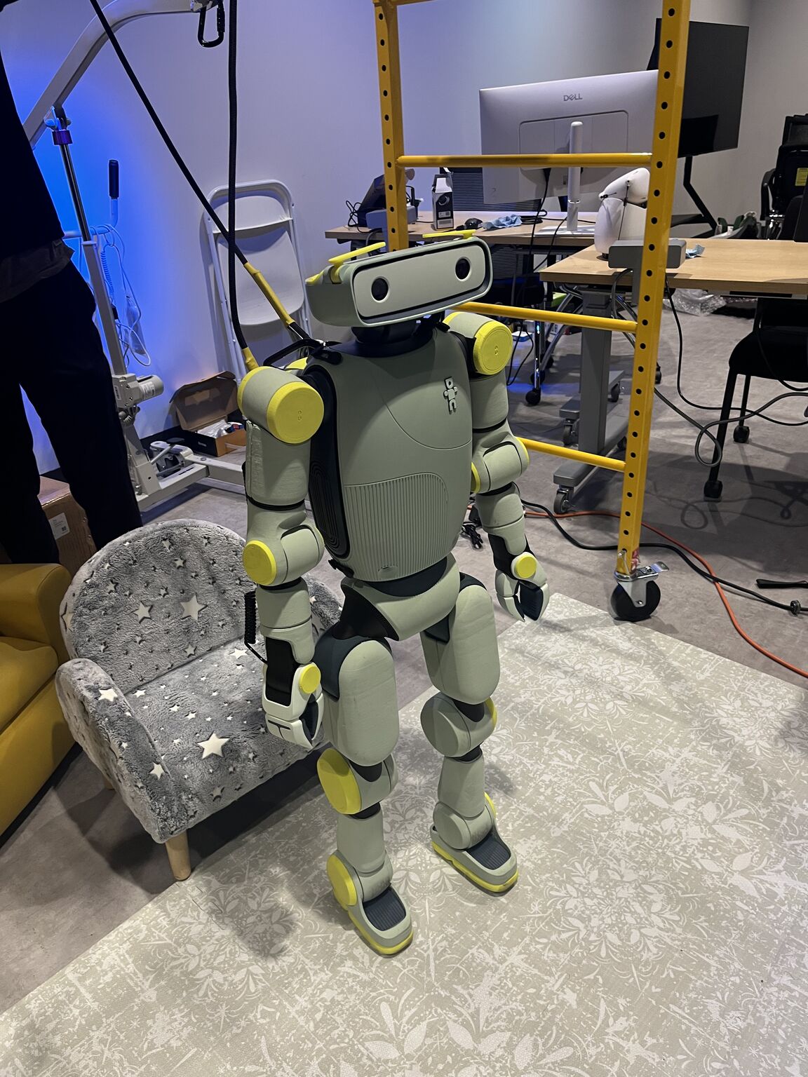 A gray humanoid robot with yellow joints stands beside a small chair and laboratory equipment.