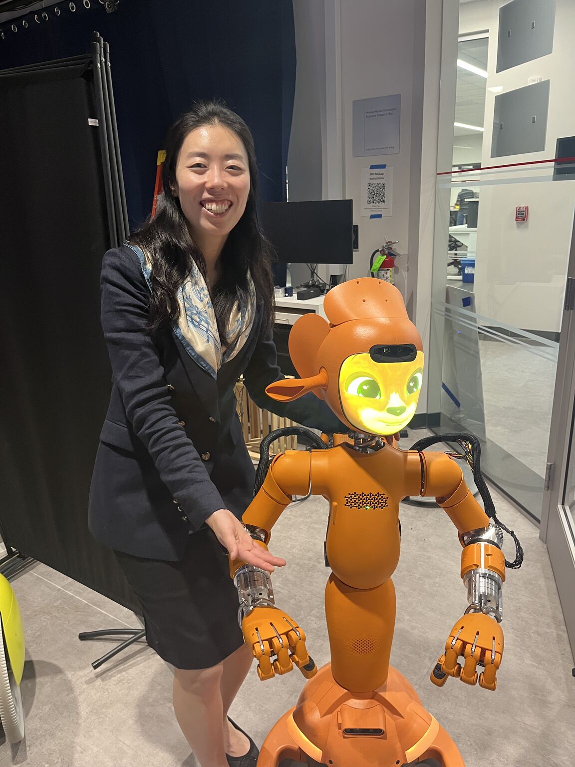A visitor stands beside an orange robot with a bright animated face and articulated hands.