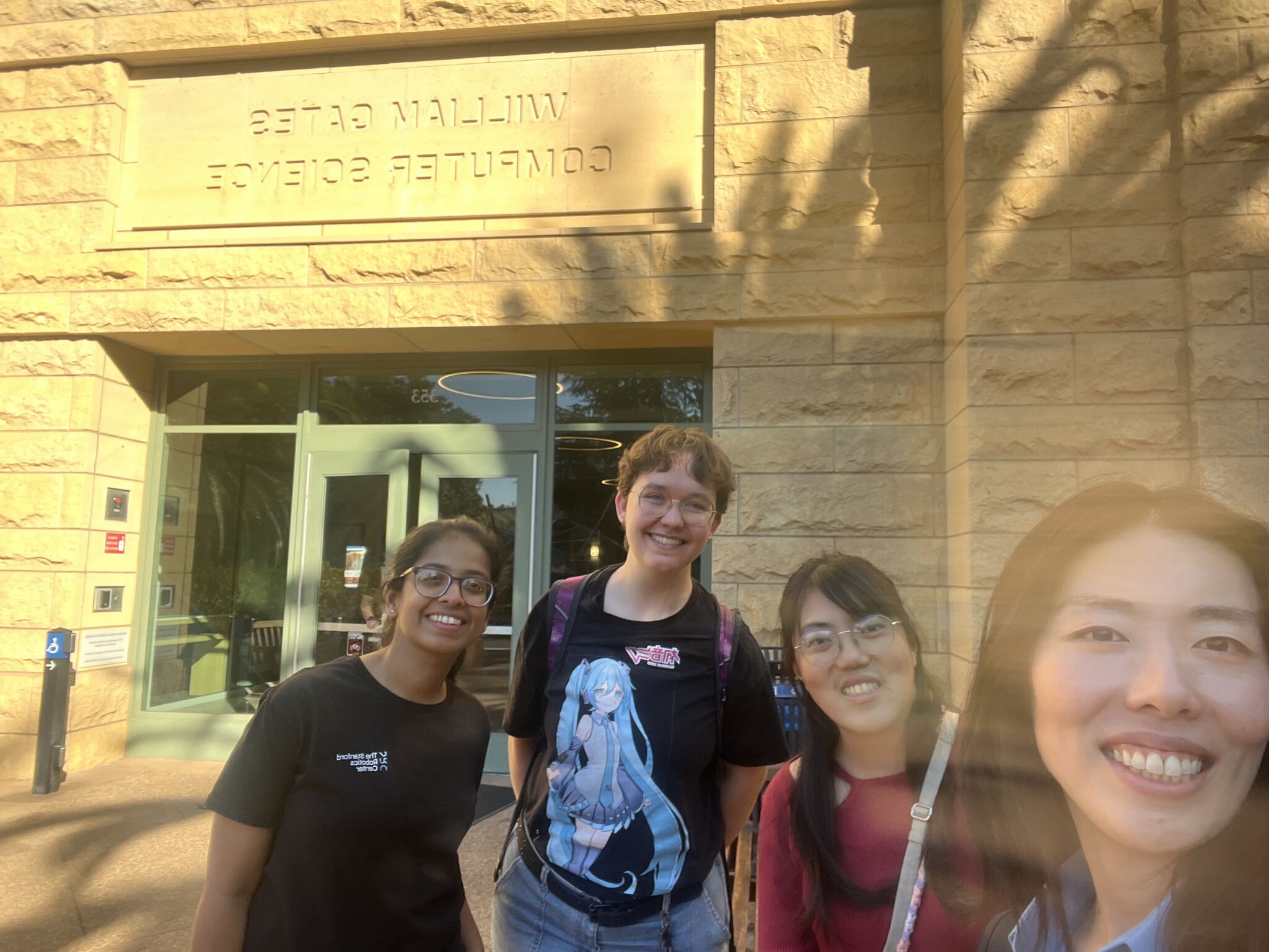 Four people pose for a selfie outside a stone building with mirrored William Gates Computer Science lettering.